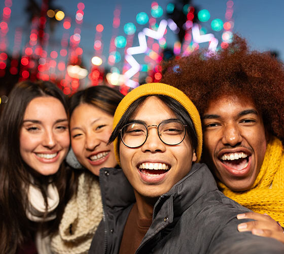 Multiracial young friends laughing and having fun together taking vertical selfie during winter market at night looking at camera. Friendship concept.